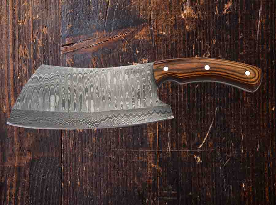 A CARLIN Damascus Cleaver by Faneema Cutlery, featuring a razor-sharp, wide, curved Damascus steel blade and a comfortable ergonomic wooden handle with rivets, rests on a textured dark wood surface.