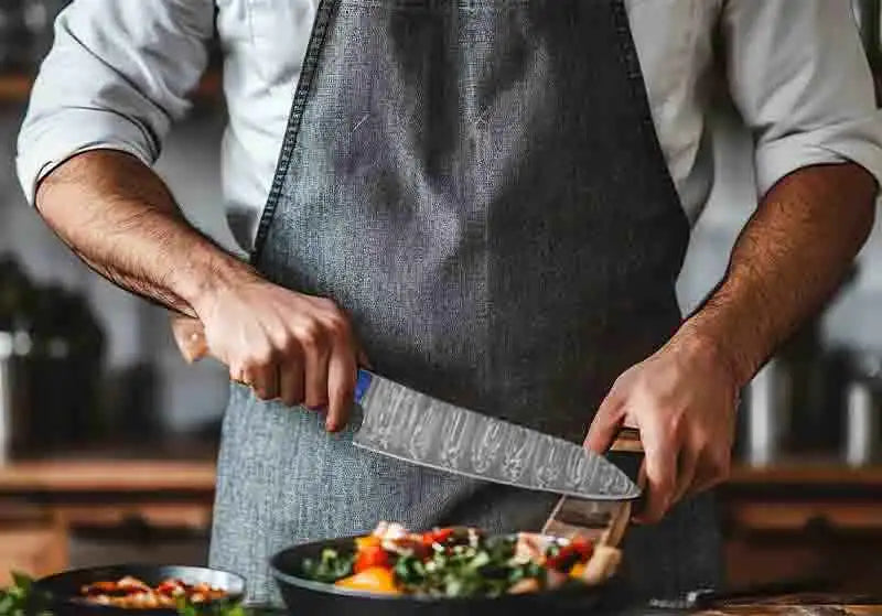 A chef’s knife with a distinctive damascus steel blade and wooden handle, worn by someone in a denim apron.