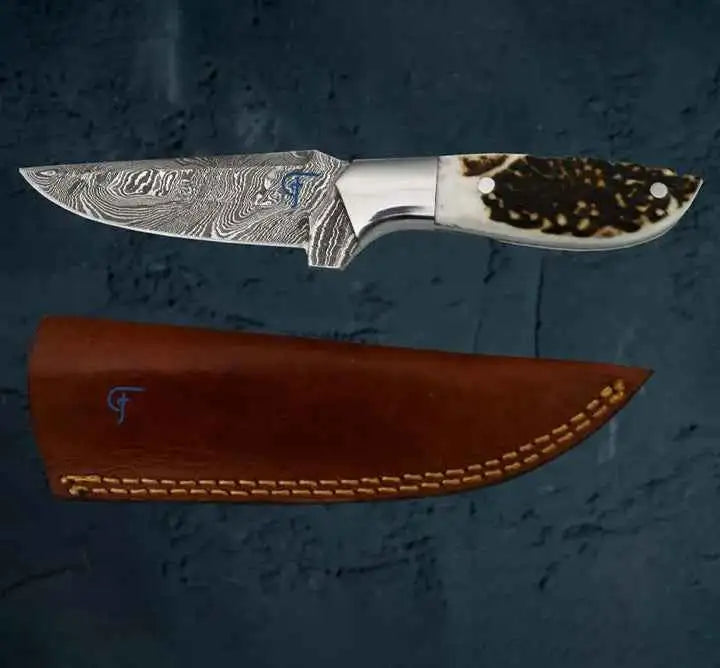 A damascus steel knife with a swirling silver blade and a white, brown-speckled antler handle rests beside its rich brown leather sheath.